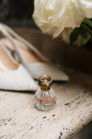 A small vintage perfume bottle sits beside white heels and roses on a distressed white surface.