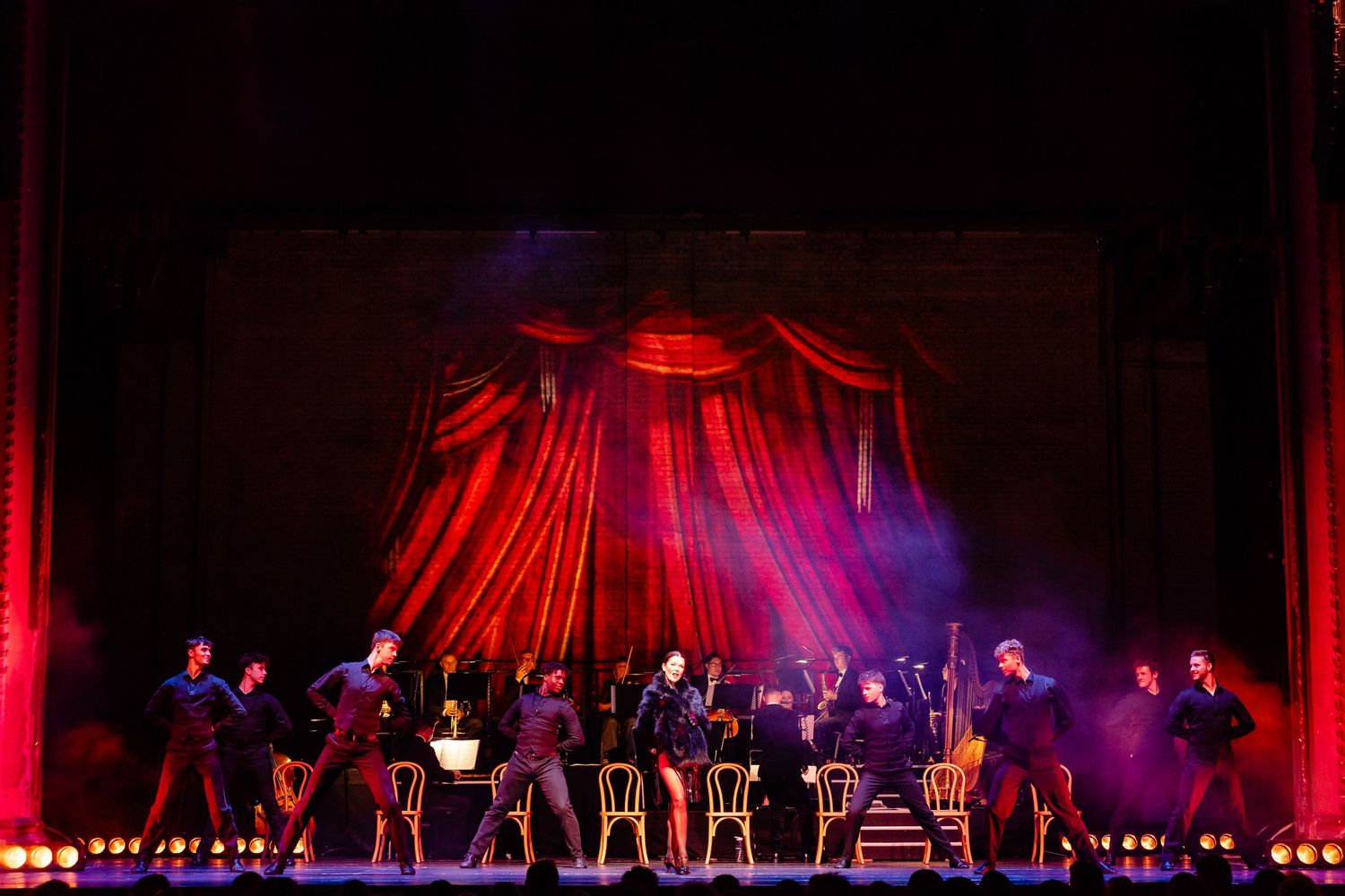 Theater ensemble performing under dramatic red curtain and lighting effects on stage.