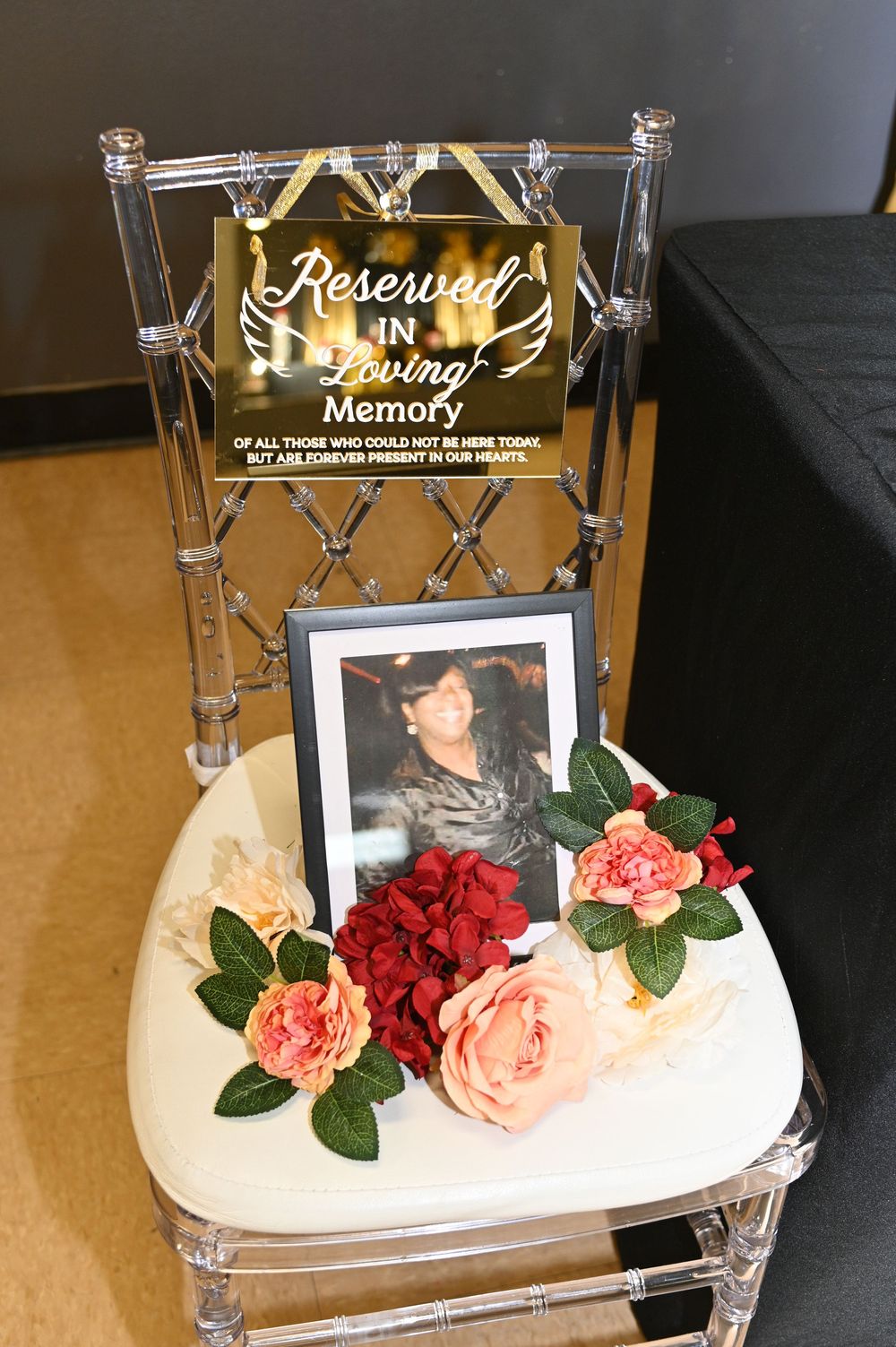 Clear acrylic chair with reserved sign, framed photograph, and pink and red roses arranged on white cushioned seat.