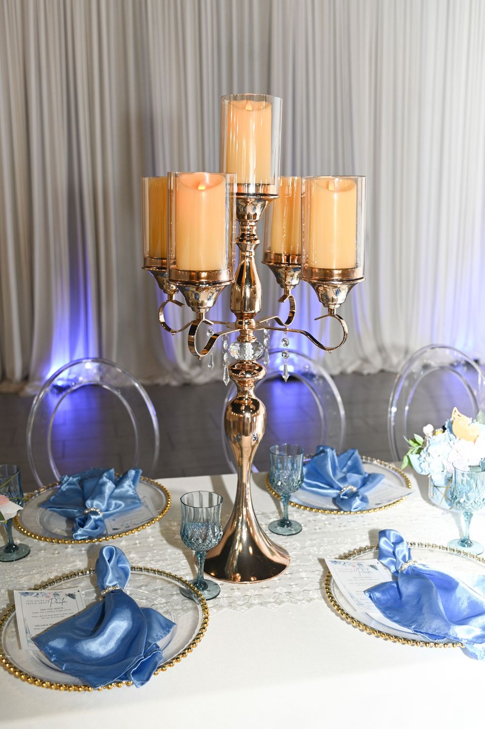 Elegant table setting with gold candelabras and blue napkins arranged on white tablecloths with decorative gold charger plates.