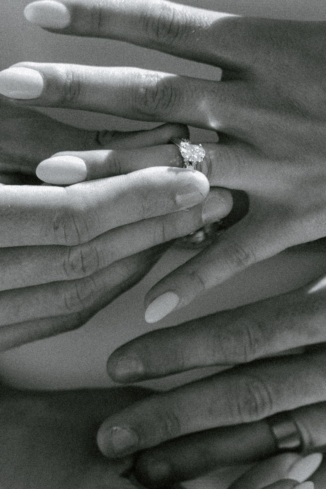 A black and white closeup of hands showcasing an elegant solitaire diamond engagement ring with manicured nails.