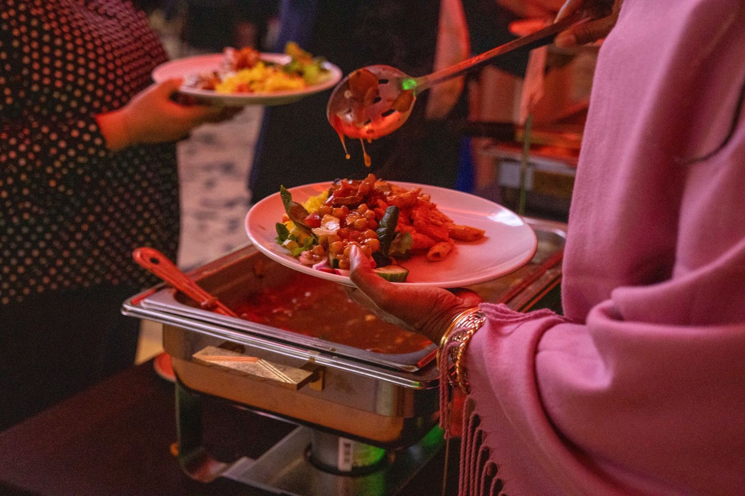 Hands serving plates with bright red sauce and food at a social gathering.