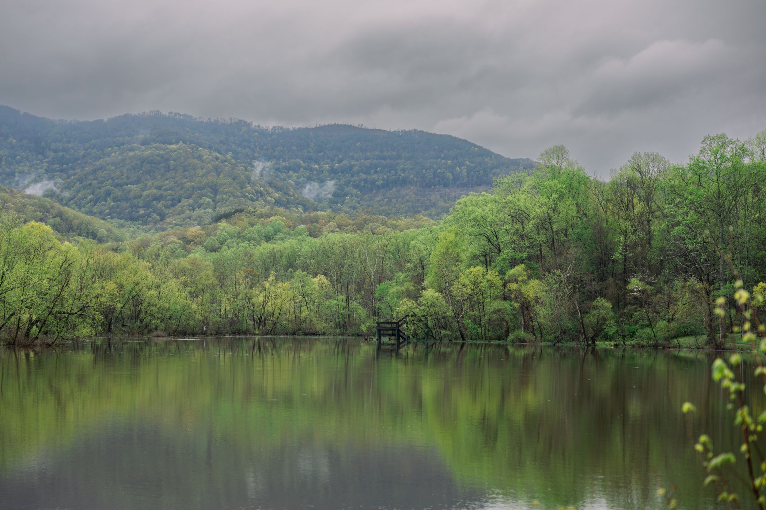 Serene mountain lake reflects lush green spring forest and misty hills under moody cloudy skies.