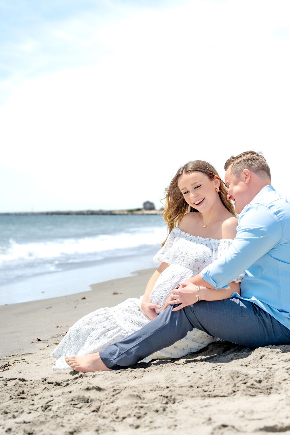 Couple sitting on sandy beach, woman in white dress laughing, man in blue shirt, ocean waves in background.