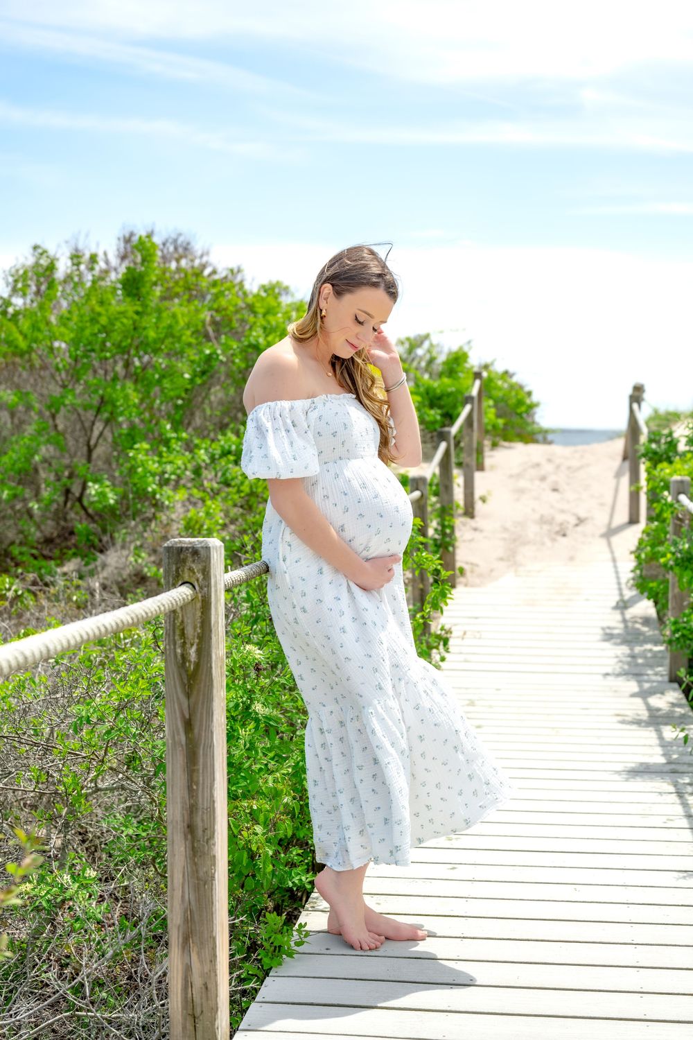 Pregnant woman in white floral off-shoulder dress standing on wooden boardwalk path surrounded by green foliage near beach.