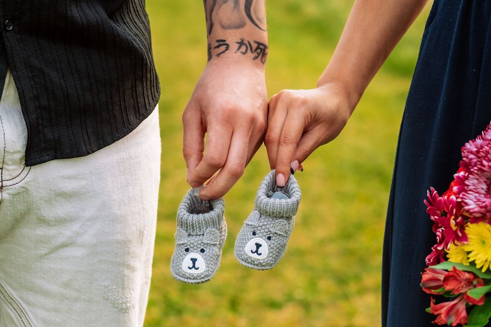 Close-up of hands holding tiny gray bear-themed baby booties against a green outdoor background.