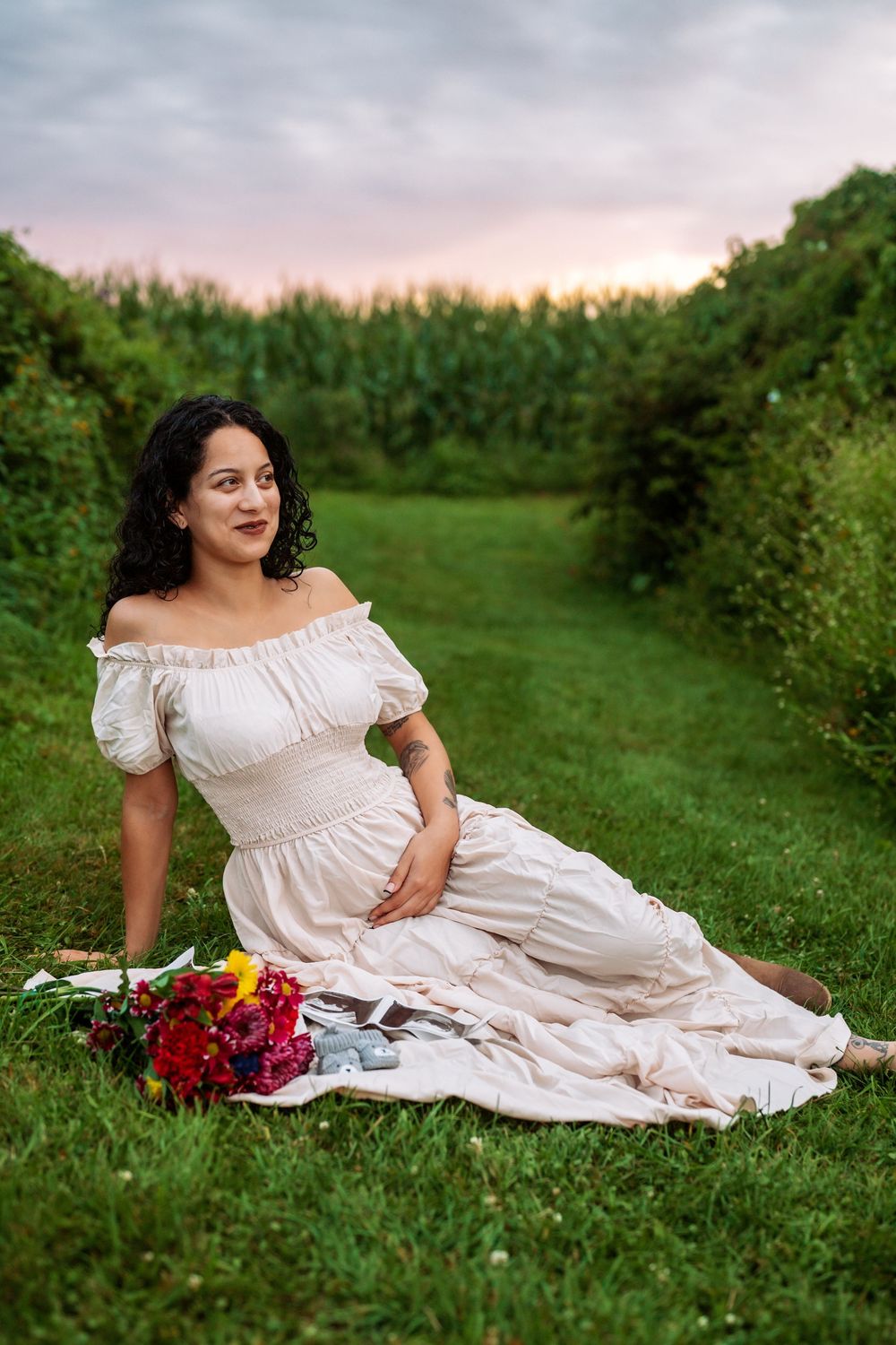 Woman in white off-shoulder dress sitting on blanket with colorful bouquet in garden setting.
