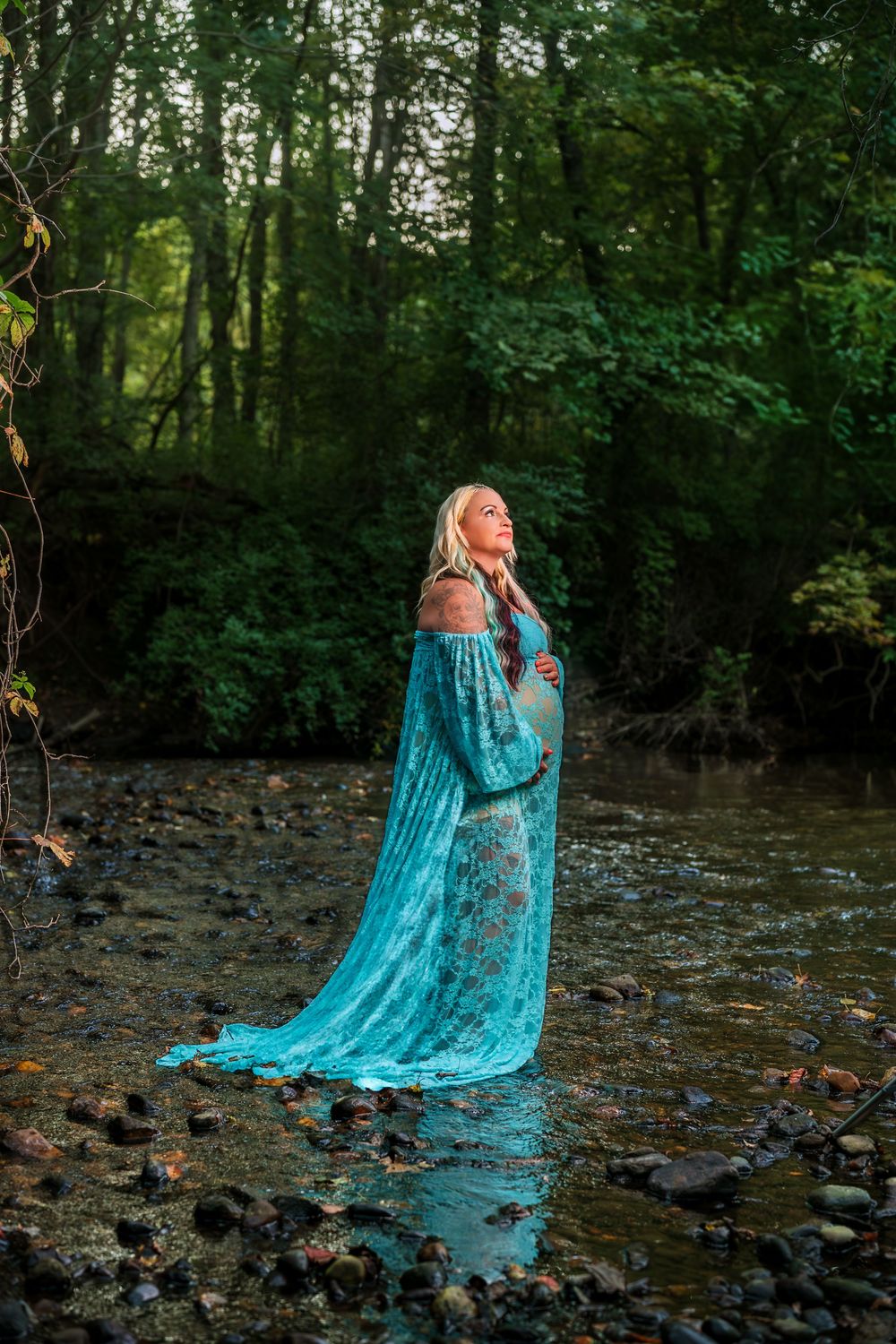Person wearing turquoise flowing dress standing in forest stream at golden hour.