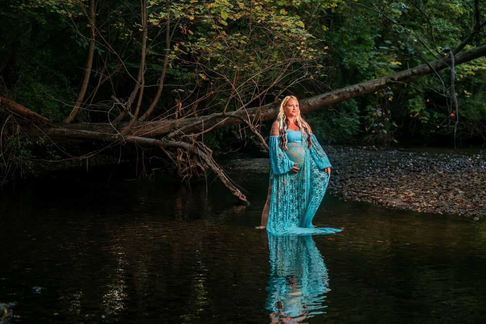 Person in flowing turquoise dress standing in forest stream with dramatic lighting.
