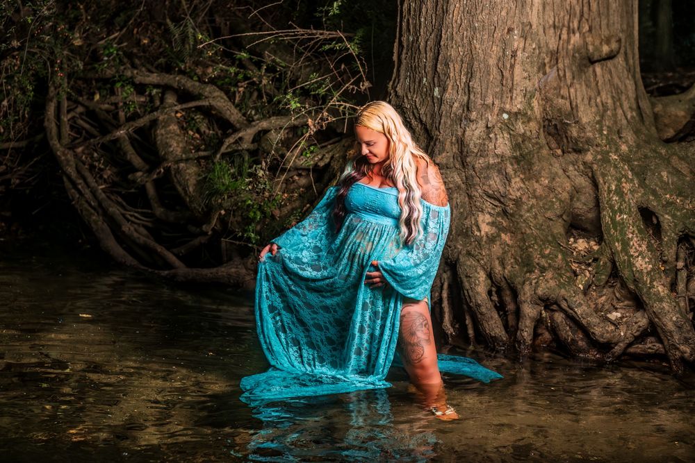 Woman in flowing turquoise dress poses artistically by a tree trunk near water.