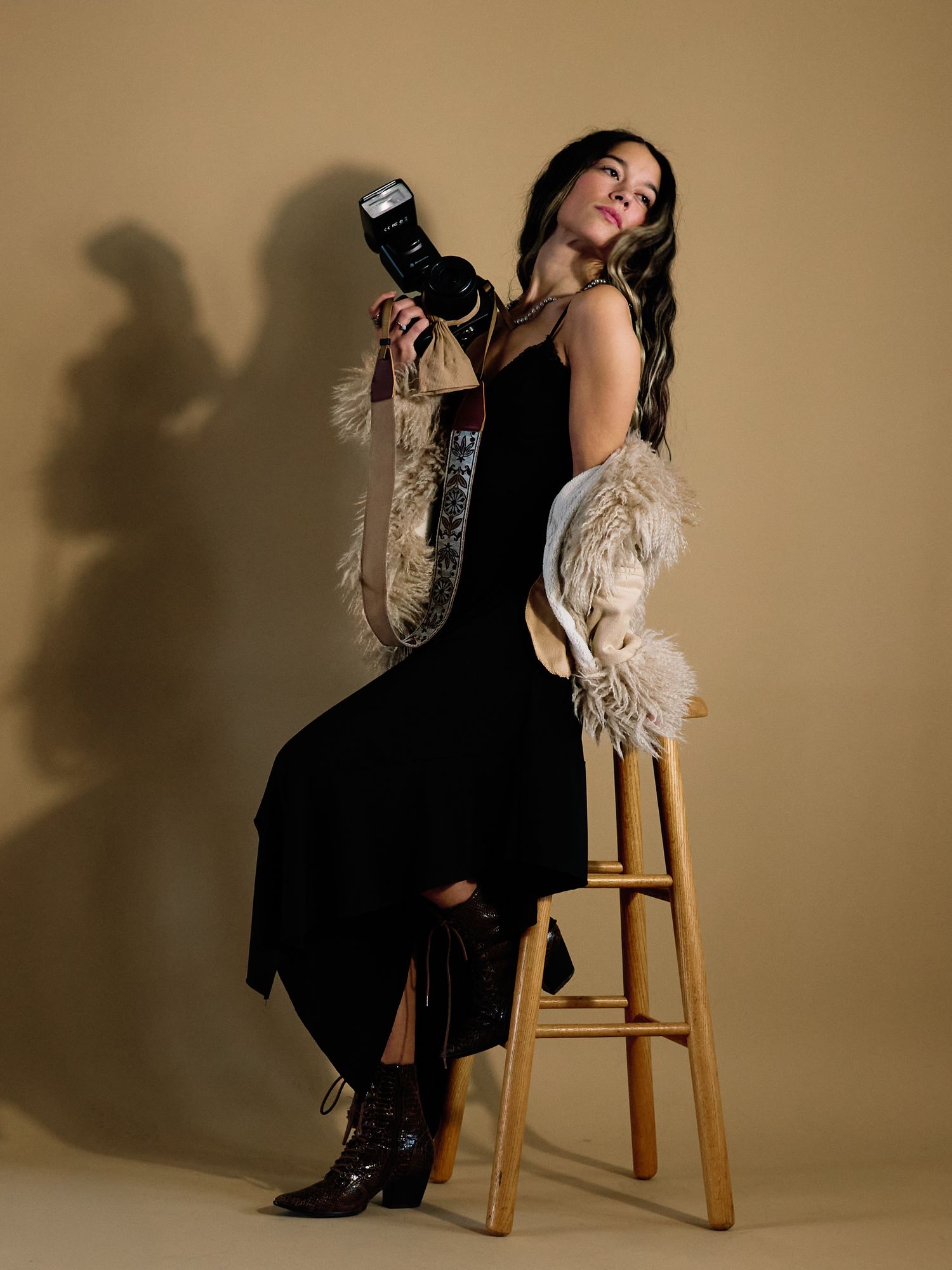 Professional studio portrait featuring a figure in black dress and cream cardigan posing dramatically on wooden stool.