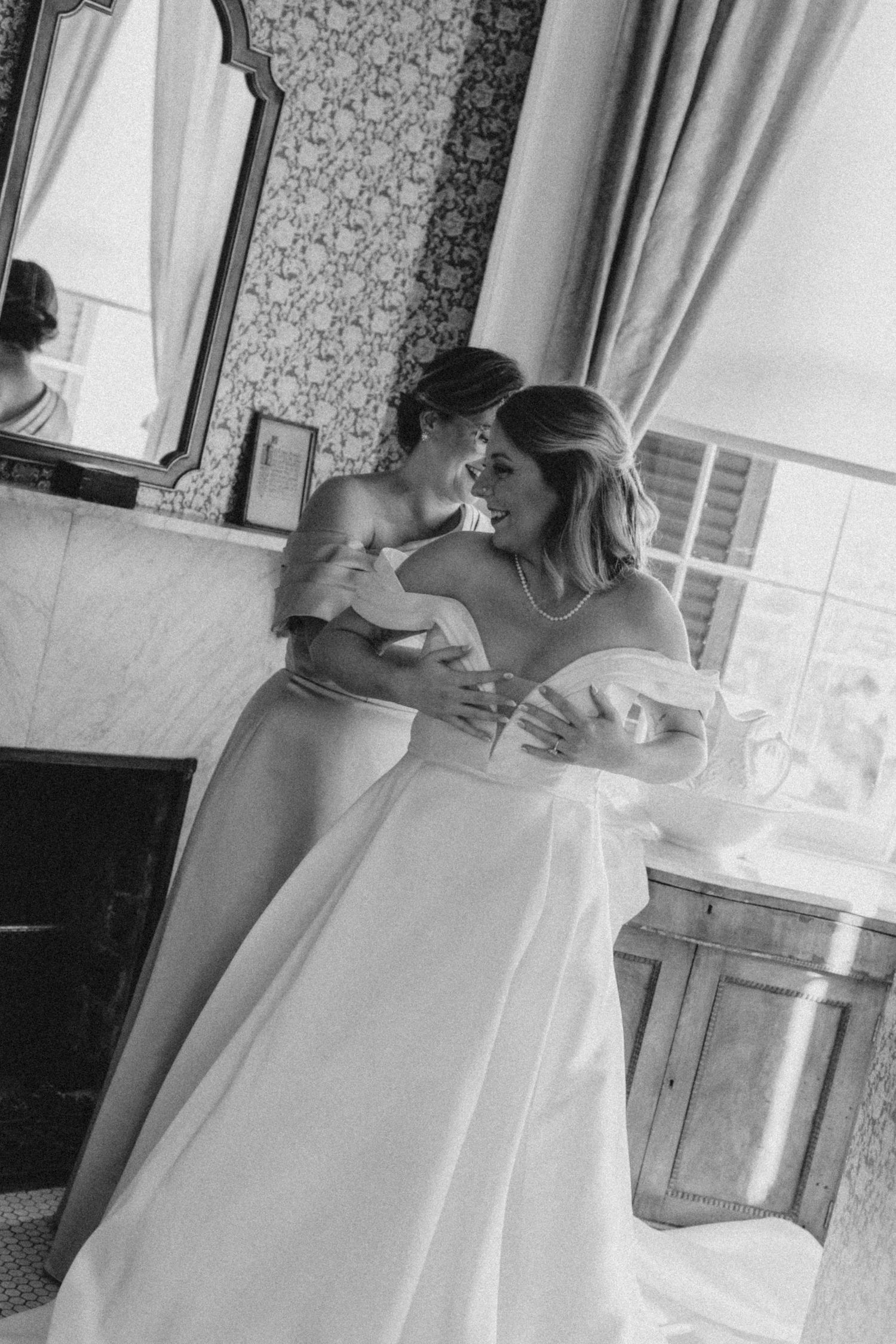 A bride holds her dress up and looks over her shoulder at her mother, standing behind her, buttoning it up