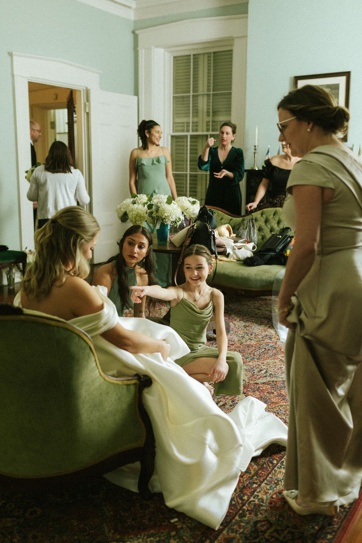 In a room full of women, a young girl kneels at the feet of a bride and points at a spot on the bride's dress while smiling