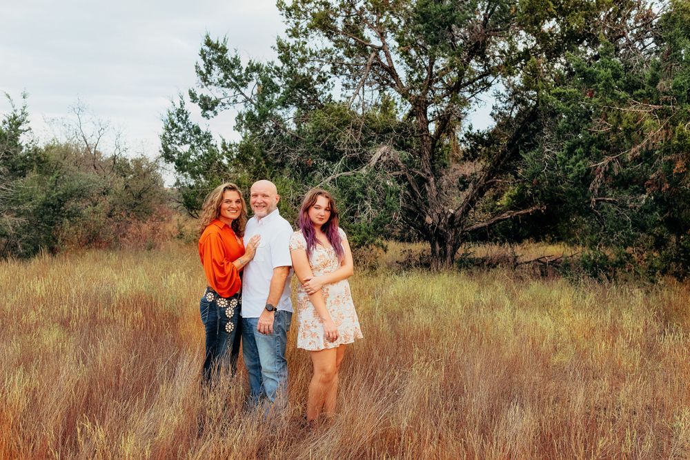 Three people stand together in a grassy field with trees behind them on an autumn day.