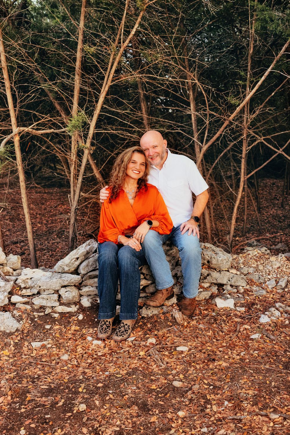 A couple sits together on a stone wall surrounded by bare tree branches in an autumn woodland setting.