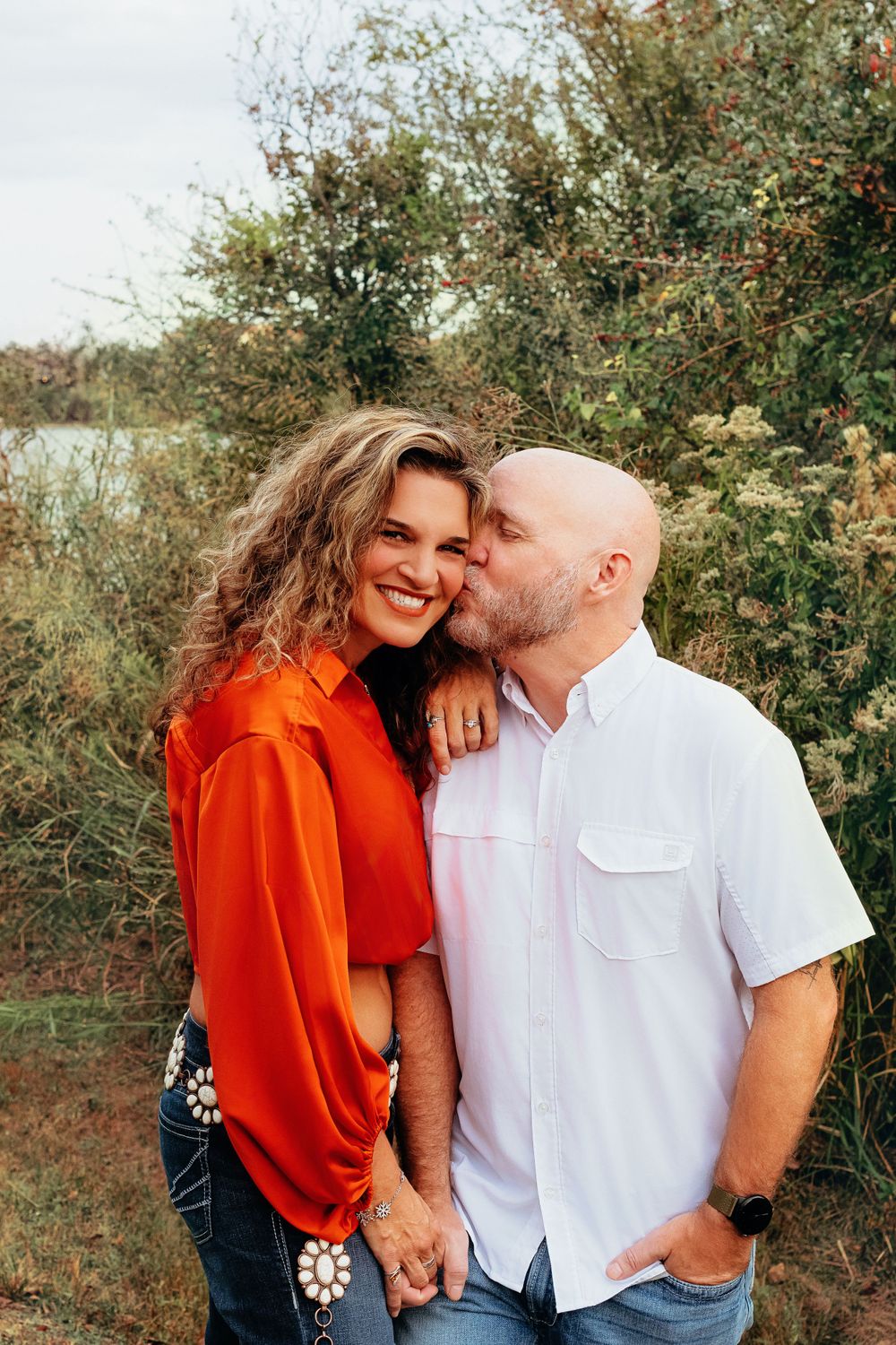 Couple embraces outdoors in front of greenery, with one wearing bright orange top and the other in white shirt.