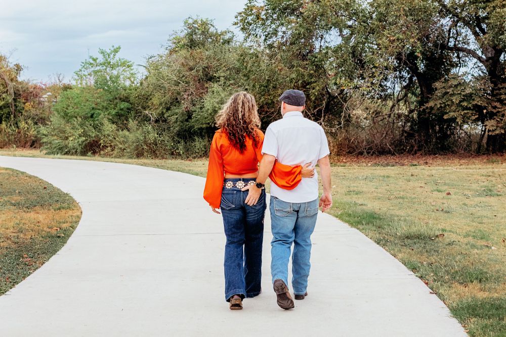 Two people walk together holding hands on a winding paved path through a grassy park on a sunny day.