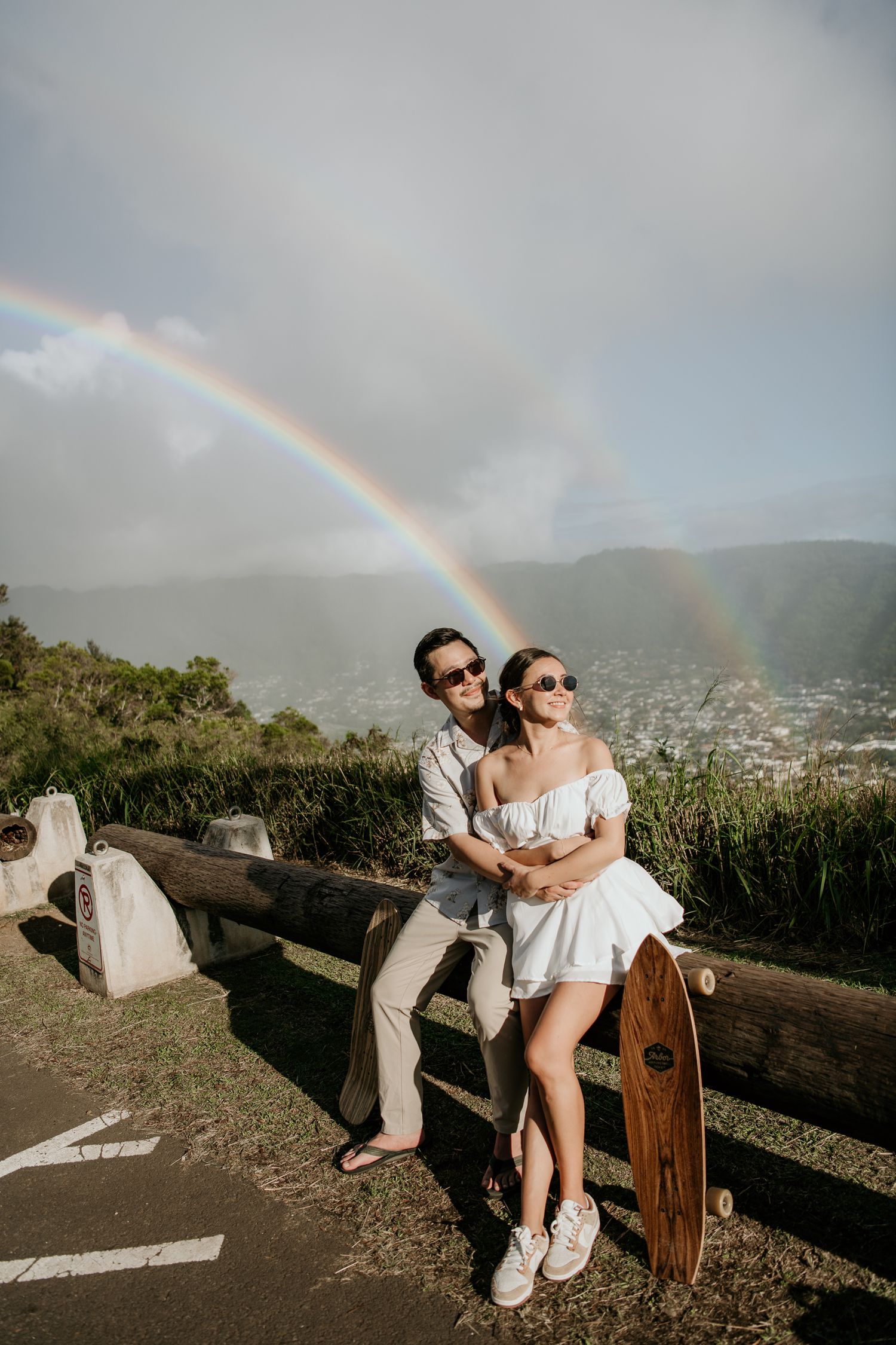 A couple stands together on a scenic overlook with a double rainbow in the sky and mountains in the background.