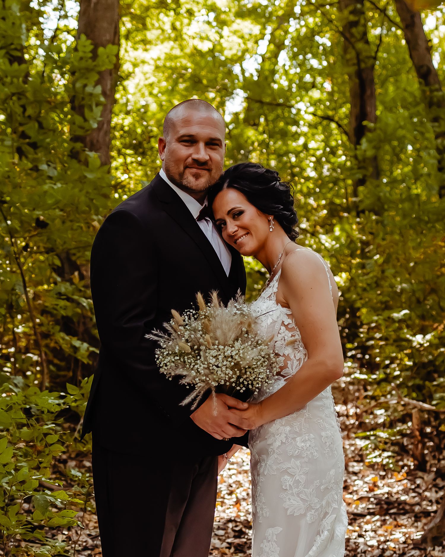 A couple embraces in a sun-dappled forest setting while holding a delicate white flower bouquet at their wedding.