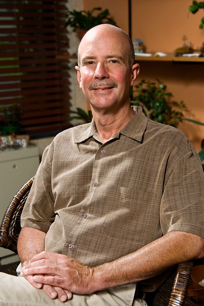 A bald man with a mustache wearing a tan plaid polo shirt sits smiling in a rattan chair against a warm interior background. (Sweden NY)