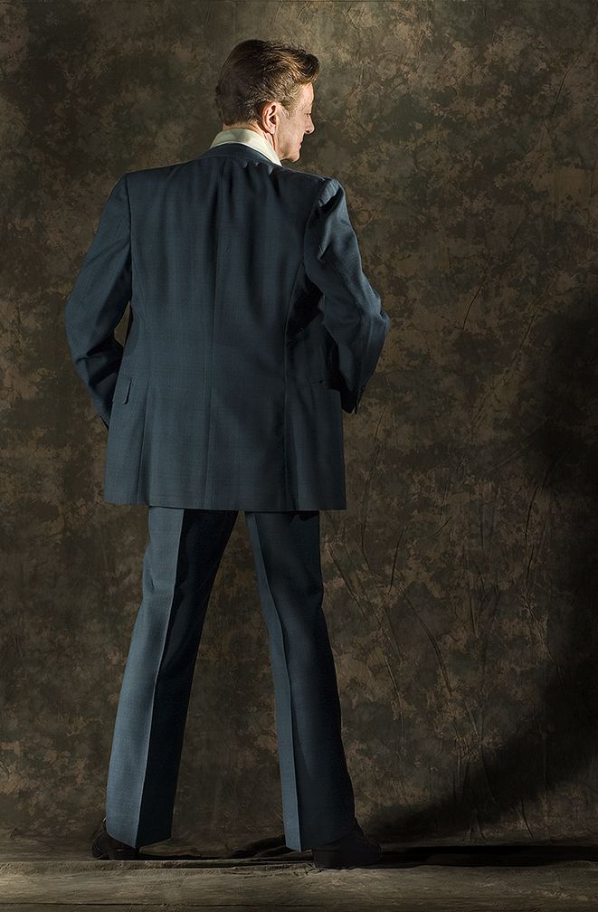 A man in a dark business suit stands with his back to the camera against a textured brown background. (Brockport New York)
