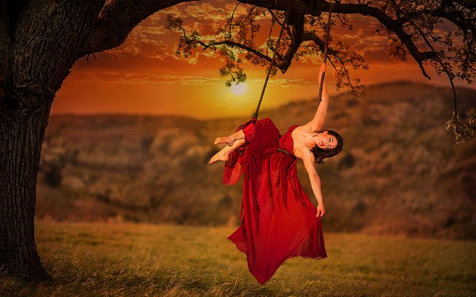 A woman in a flowing red dress swings gracefully from a tree branch against a dramatic orange sunset backdrop.