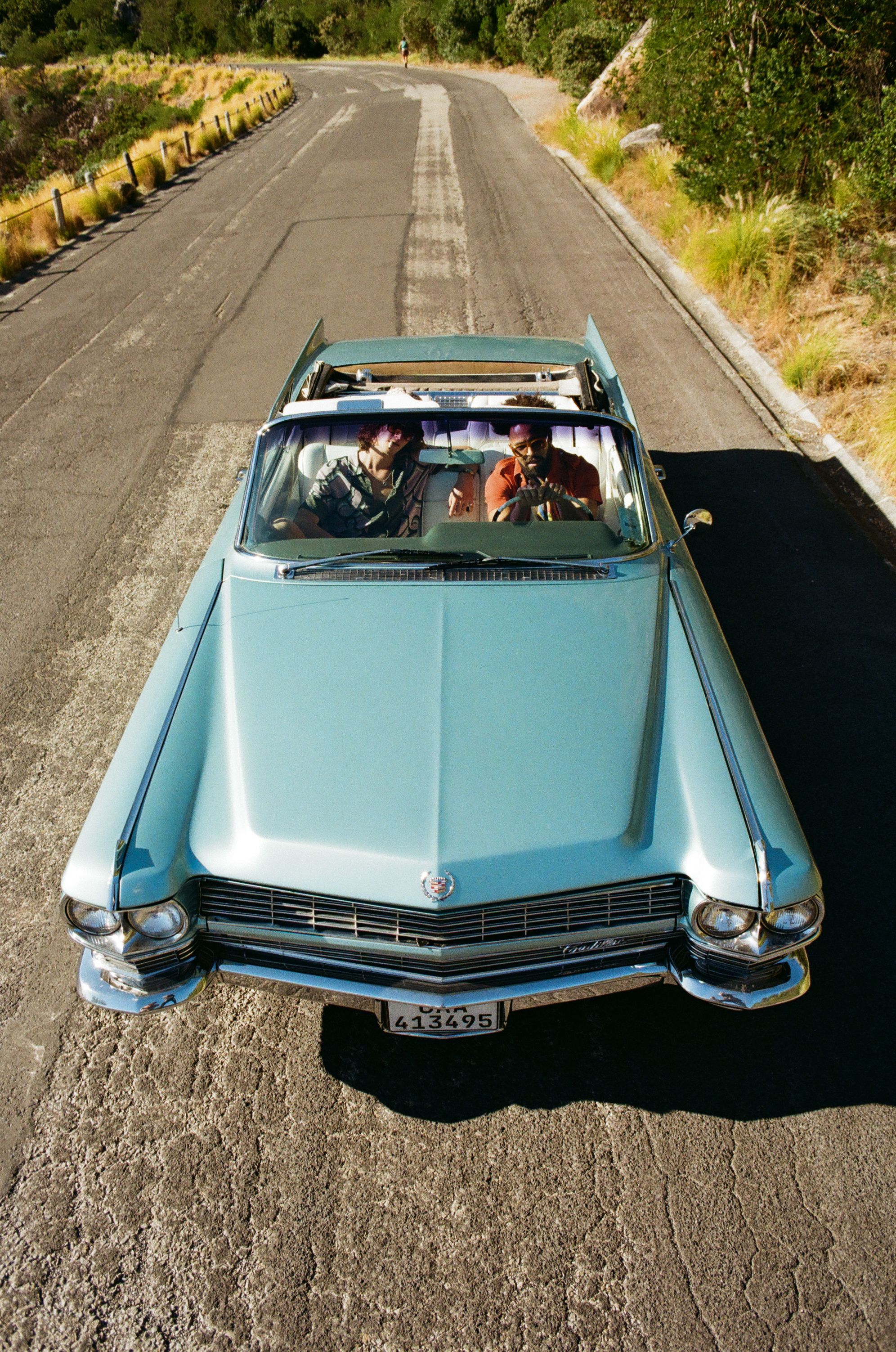 Vintage light blue Cadillac convertible driving down a winding mountain road on a sunny day.