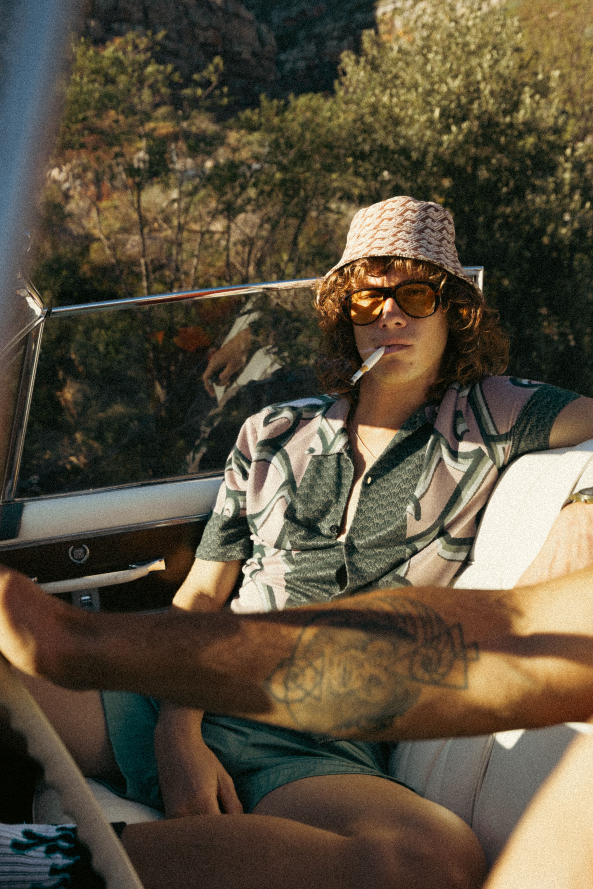 Retro-style photo of someone wearing a bucket hat and sunglasses relaxing in a car seat on a sunny day.