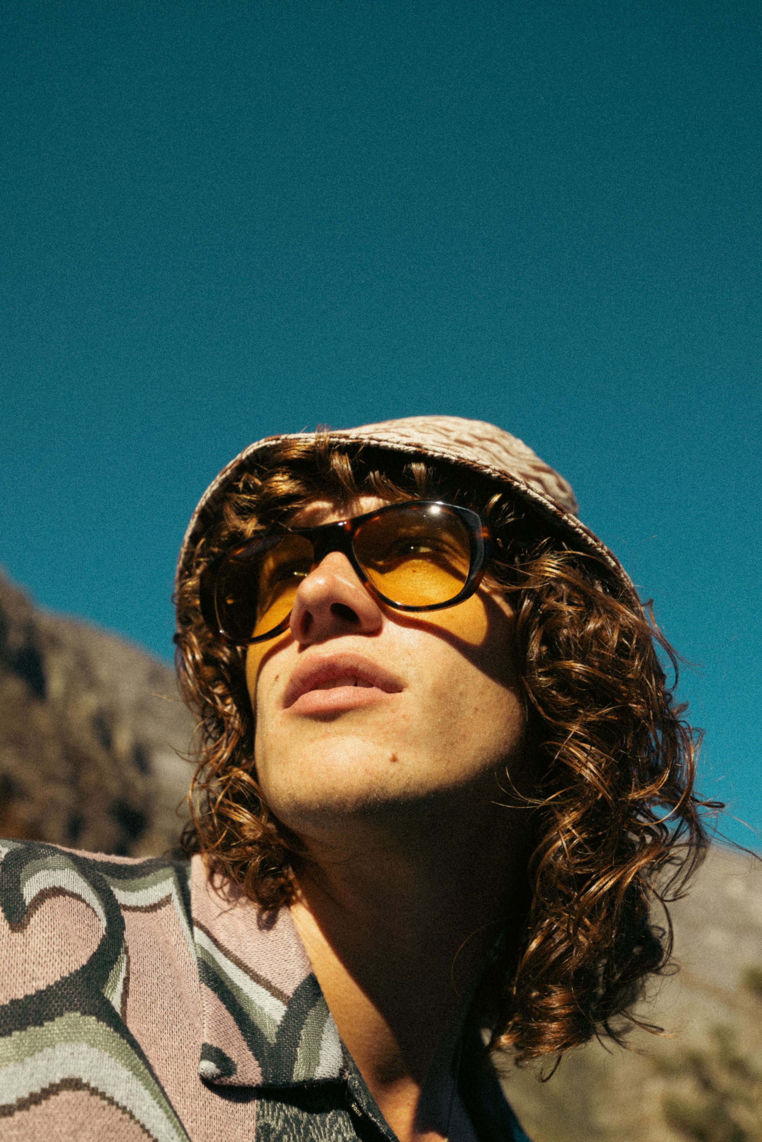 Artistic portrait of someone wearing retro sunglasses and a bucket hat against a turquoise sky backdrop.