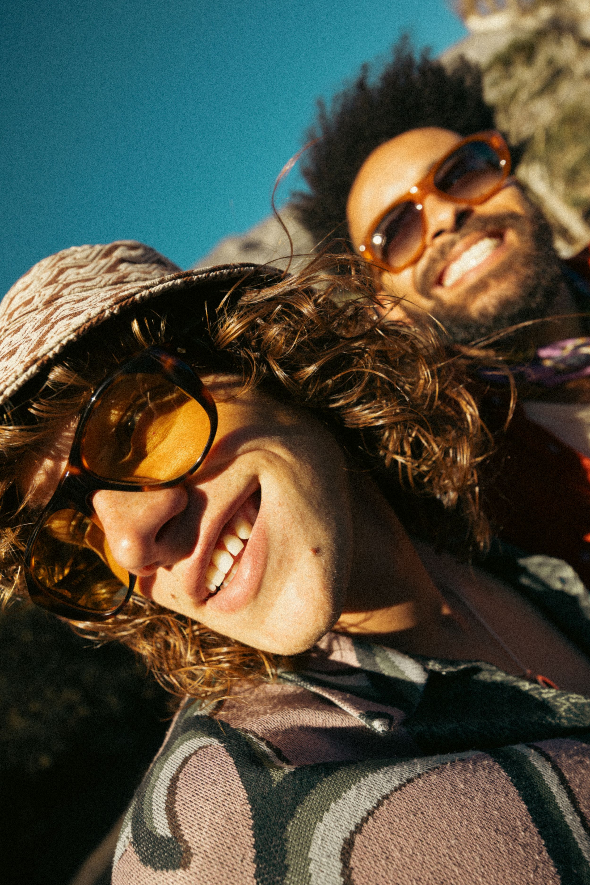 A sun-lit closeup photo of two smiling friends wearing sunglasses laying back and enjoying a sunny day outdoors.