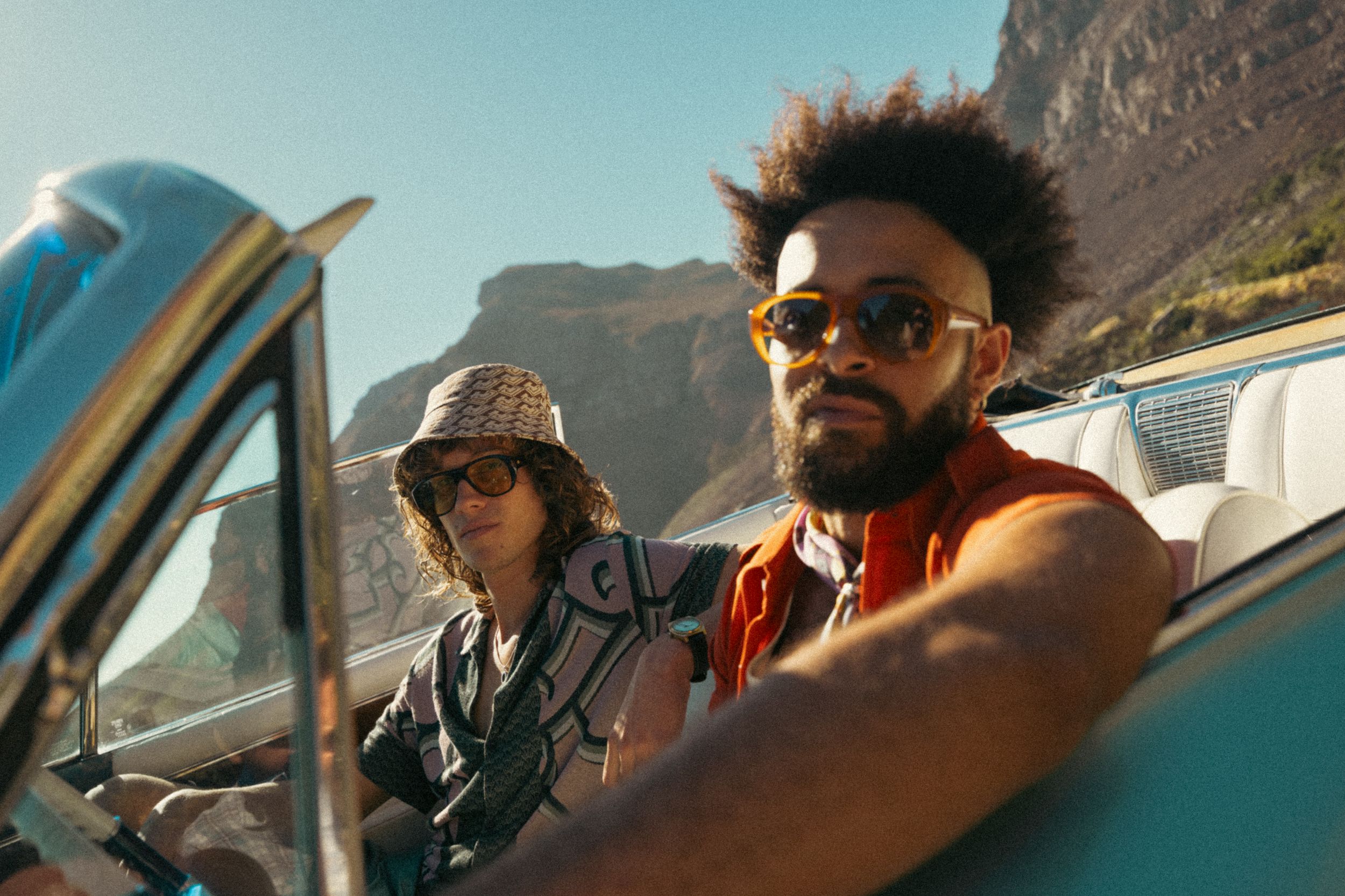 Road trip companions driving through scenic mountain landscape in convertible car during golden hour.