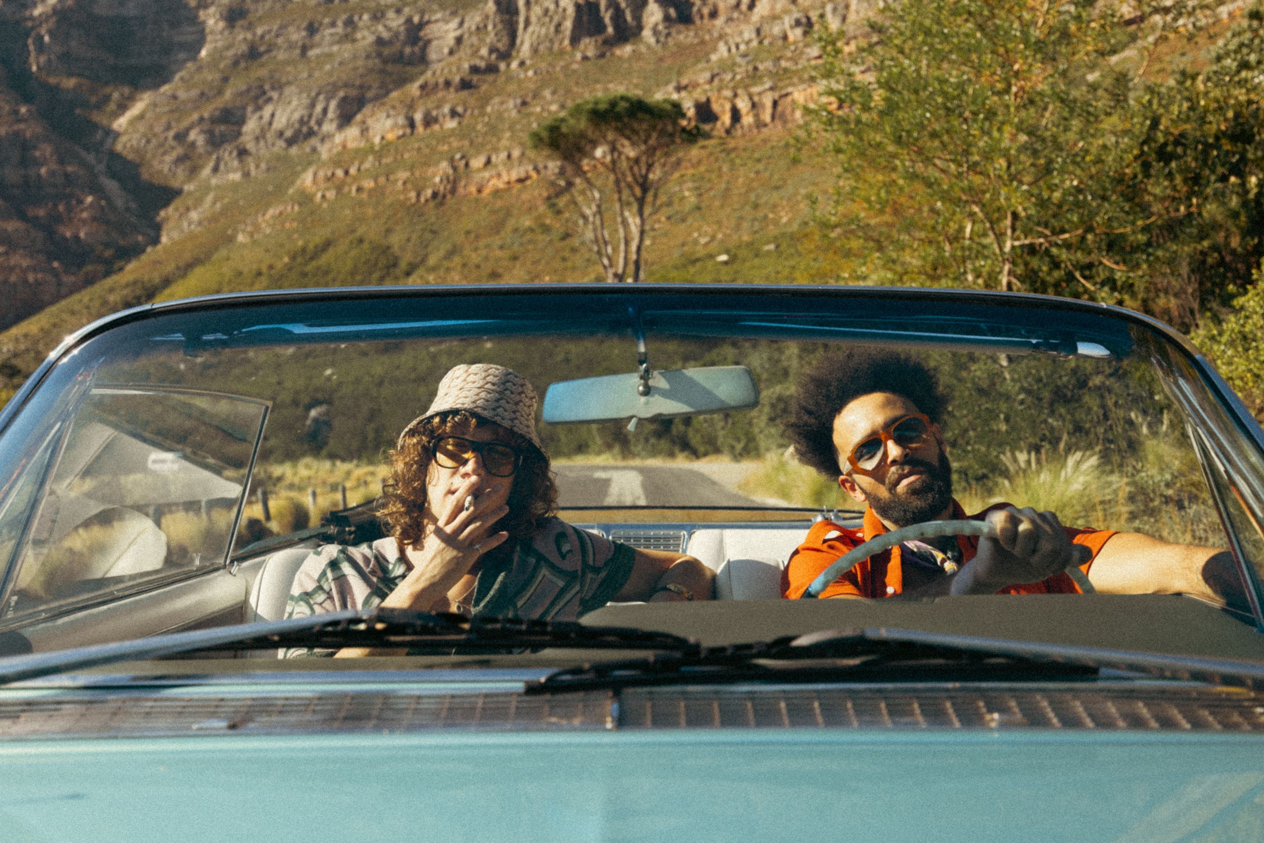 Two people sit in a convertible car with mountainous terrain visible in the background on a sunny day.