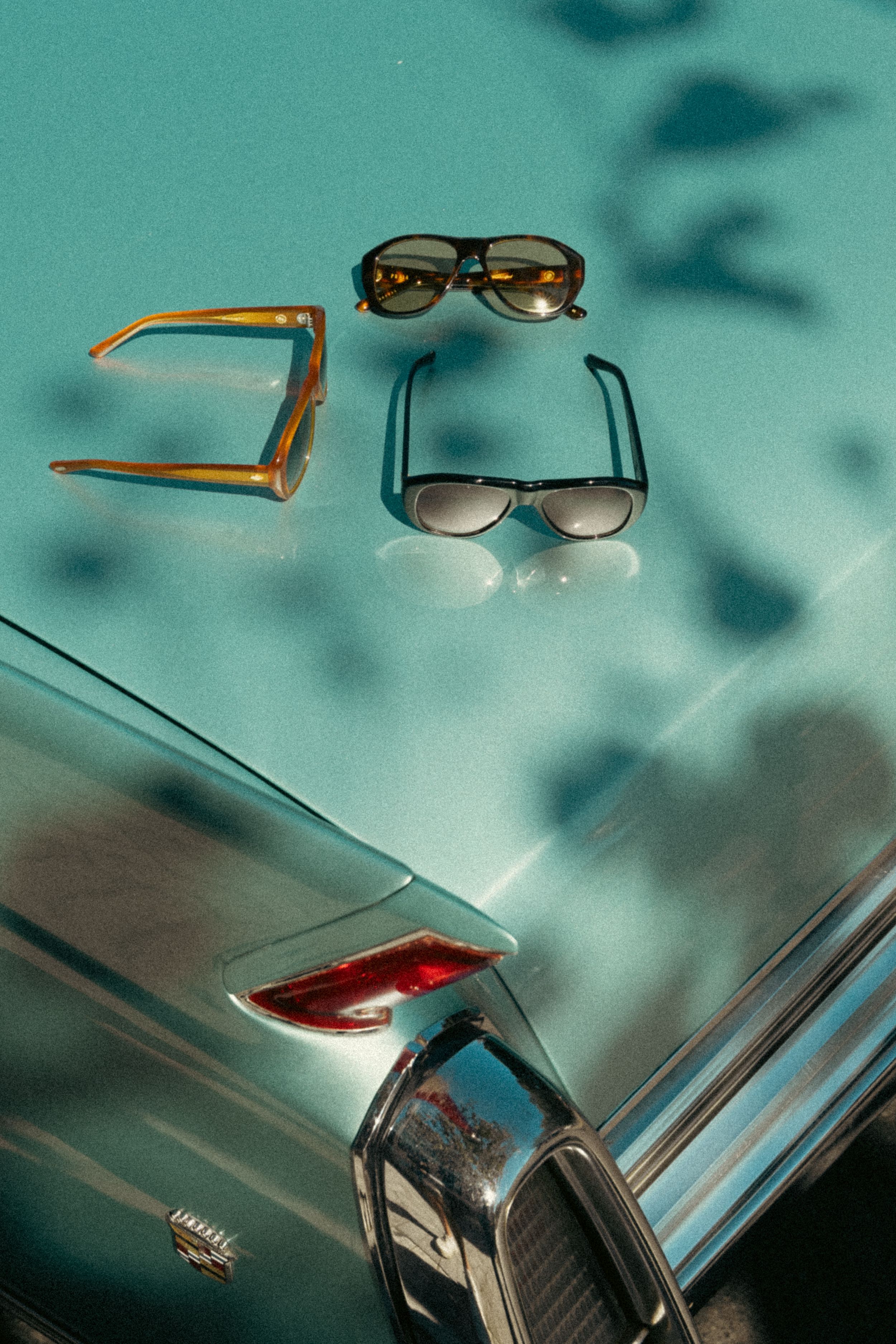 Two pairs of vintage sunglasses rest on a turquoise surface near the chrome trim of a classic car in retro styling.