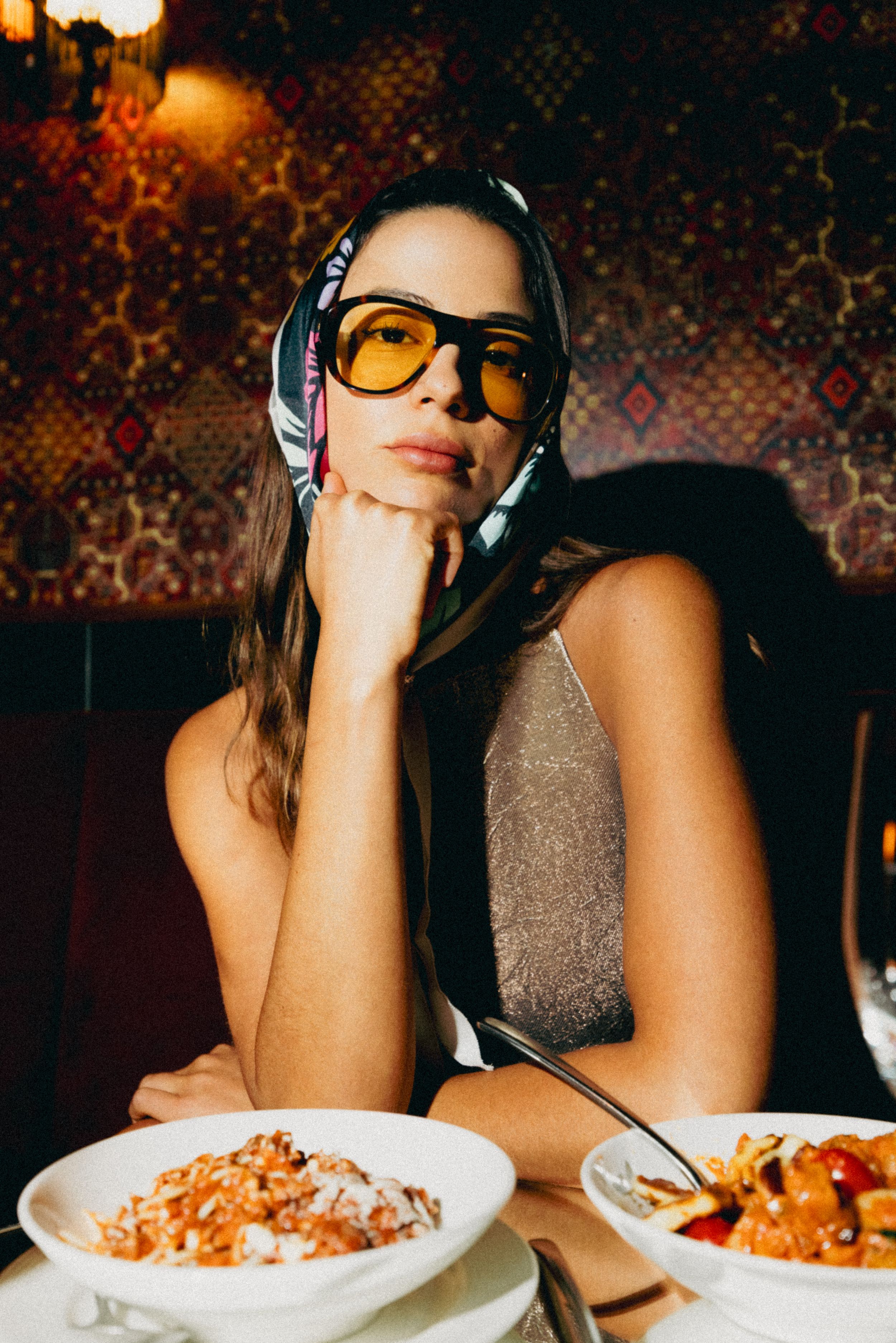 A stylish woman in yellow-tinted glasses and headscarf sits at a restaurant table with pasta dishes.