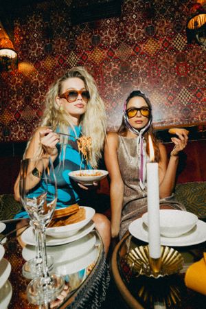 Two people in sunglasses enjoy a stylish dinner with candles and plates at a restaurant with ornate patterned walls.