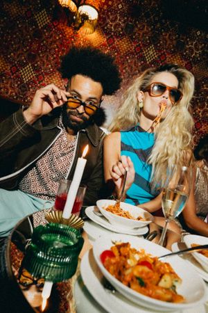 Two stylish friends enjoy a romantic candlelit dinner with pasta and wine at an intimate restaurant setting.