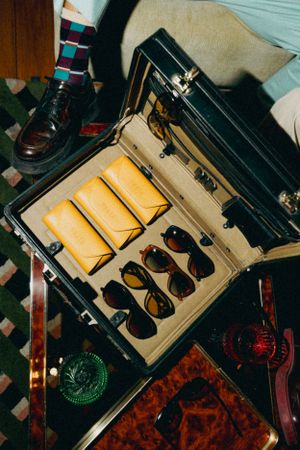 An open vintage suitcase displays a collection of retro sunglasses and yellow wallets on a patterned surface.