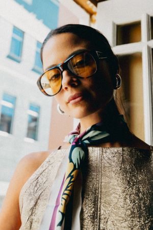 Fashion portrait of model wearing retro aviator sunglasses and patterned scarf against window light in vintage style.