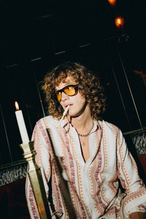 A person with curly hair wearing sunglasses and a floral patterned shirt poses with a candlestick in a dimly lit setting.