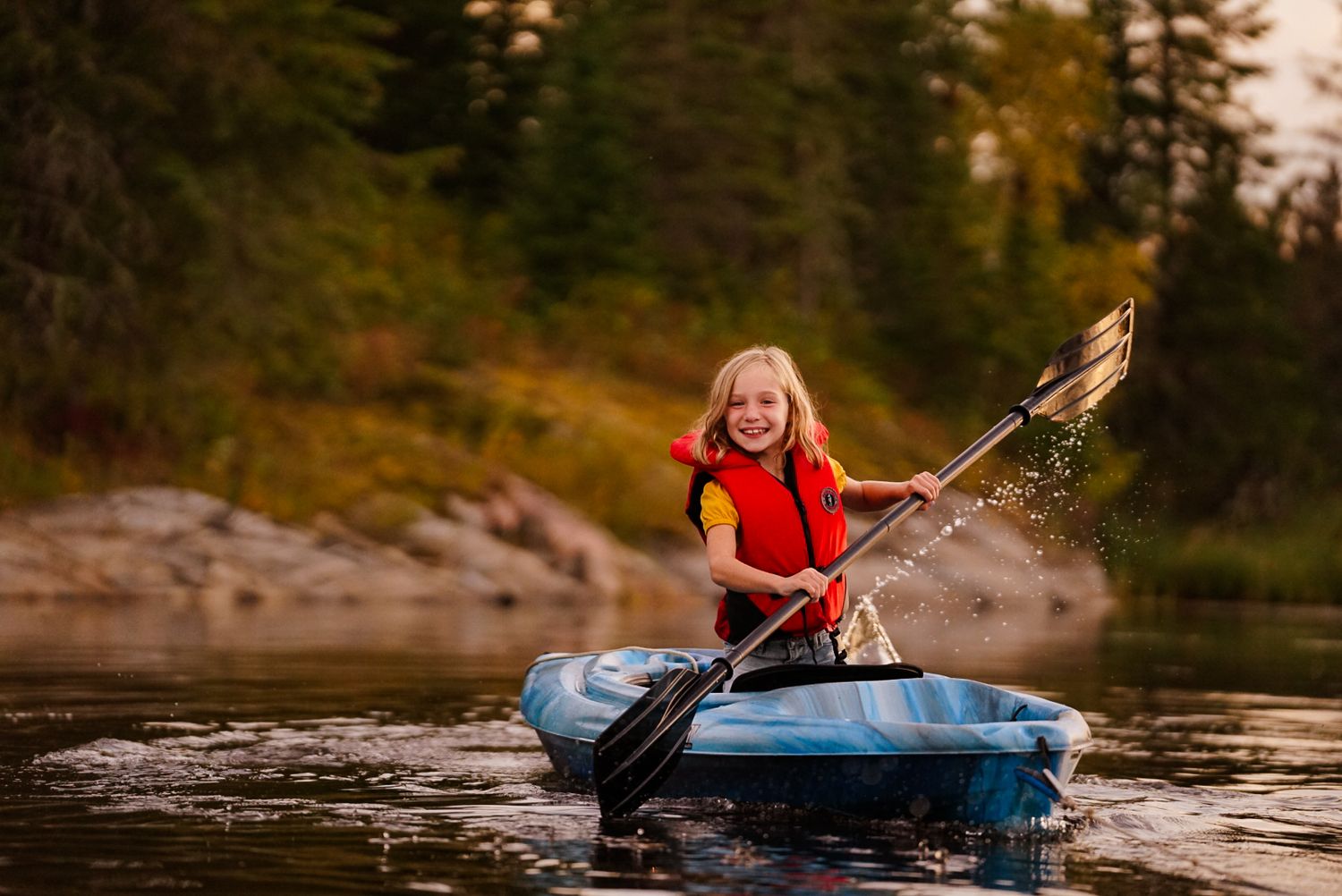 Kayakers paddle across scenic lake with rocky shoreline and evergreen trees.