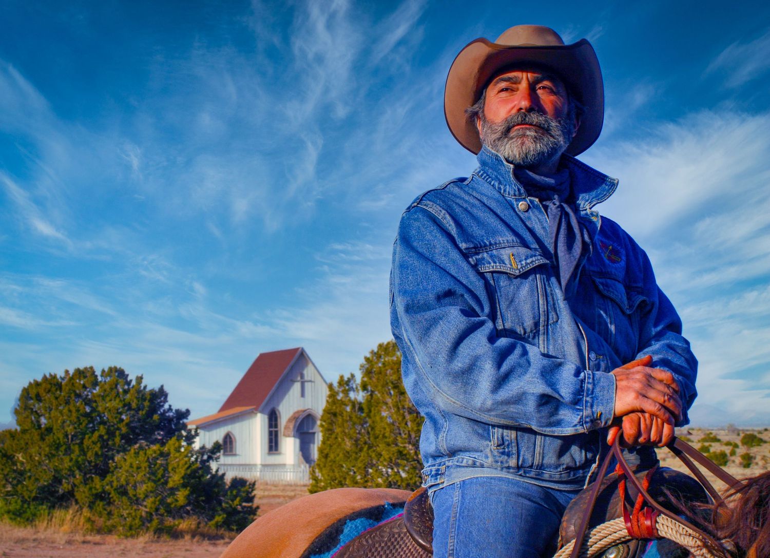 A person wearing a denim jacket and cowboy hat rides a horse through a scenic landscape under a bright blue sky.