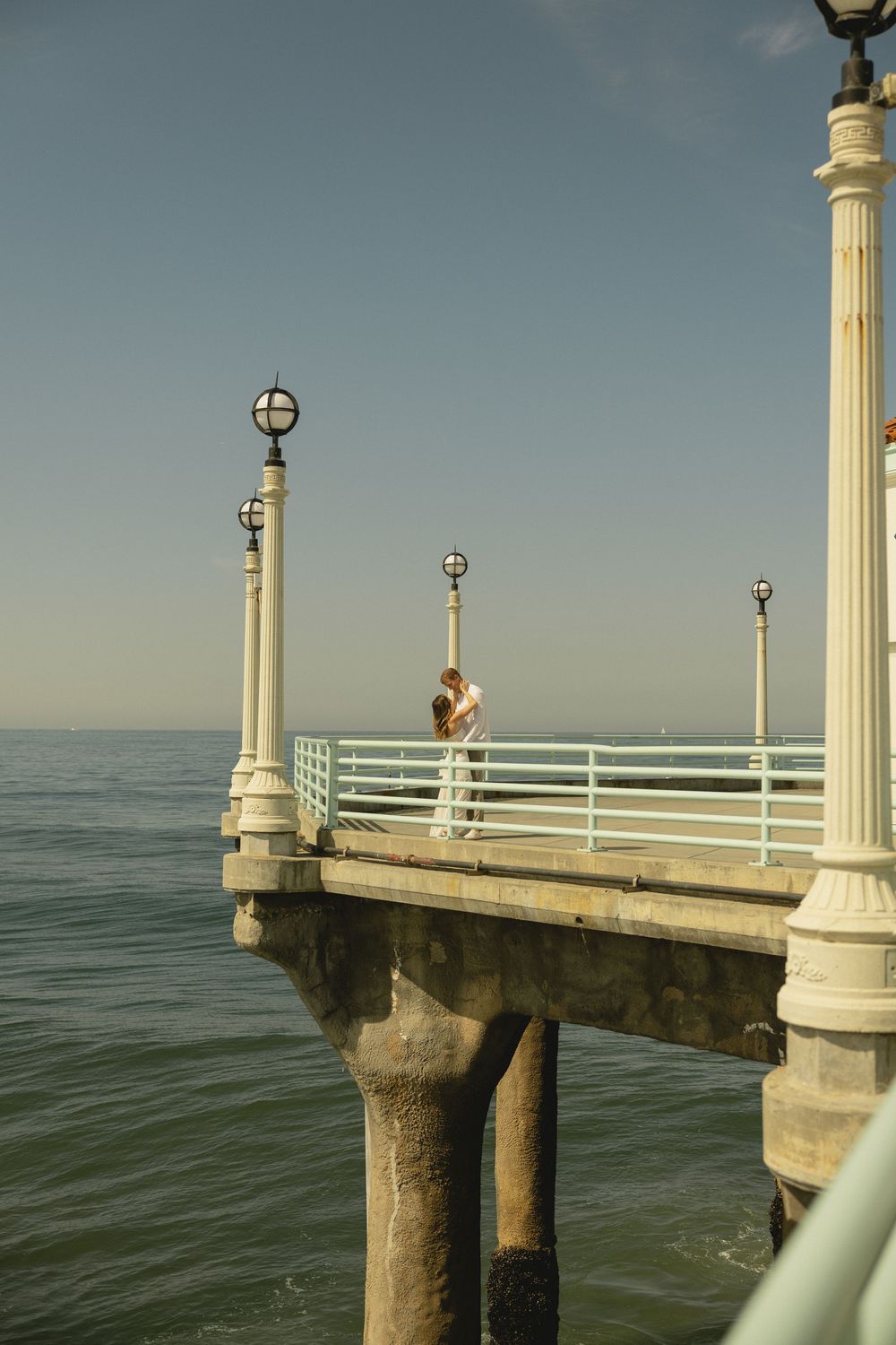 Person standing on a classic white pier with lamp posts overlooking calm ocean waters under a clear blue sky.