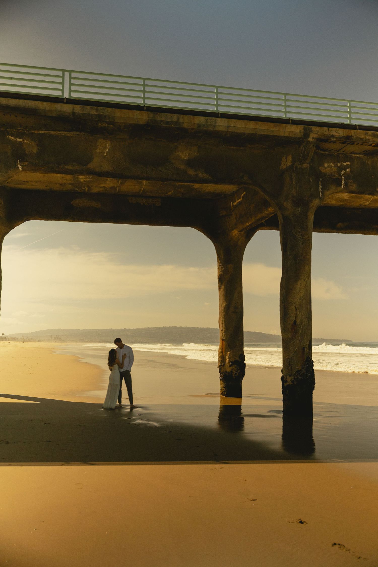 Couple embracing romantically under a concrete pier on a golden sandy beach at sunset with ocean waves in the background.