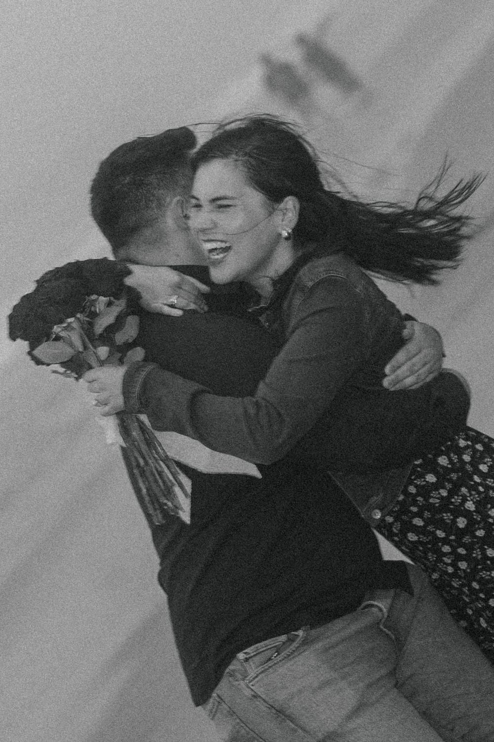 Black and white photo of a joyful couple embracing outdoors, woman laughing as her hair blows in the wind.