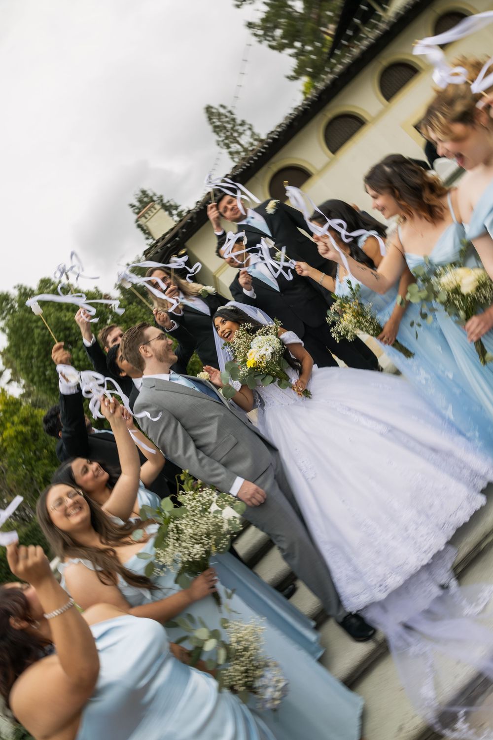 Wedding party celebrates outdoors, bride in white gown with bridesmaids in blue dresses and groomsmen waving ribbon wands.