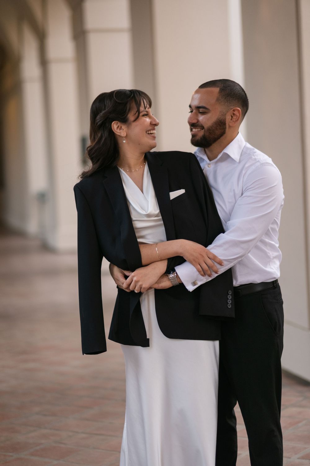 A couple in elegant black and white attire embraces and smiles at each other in a sunlit architectural hallway.