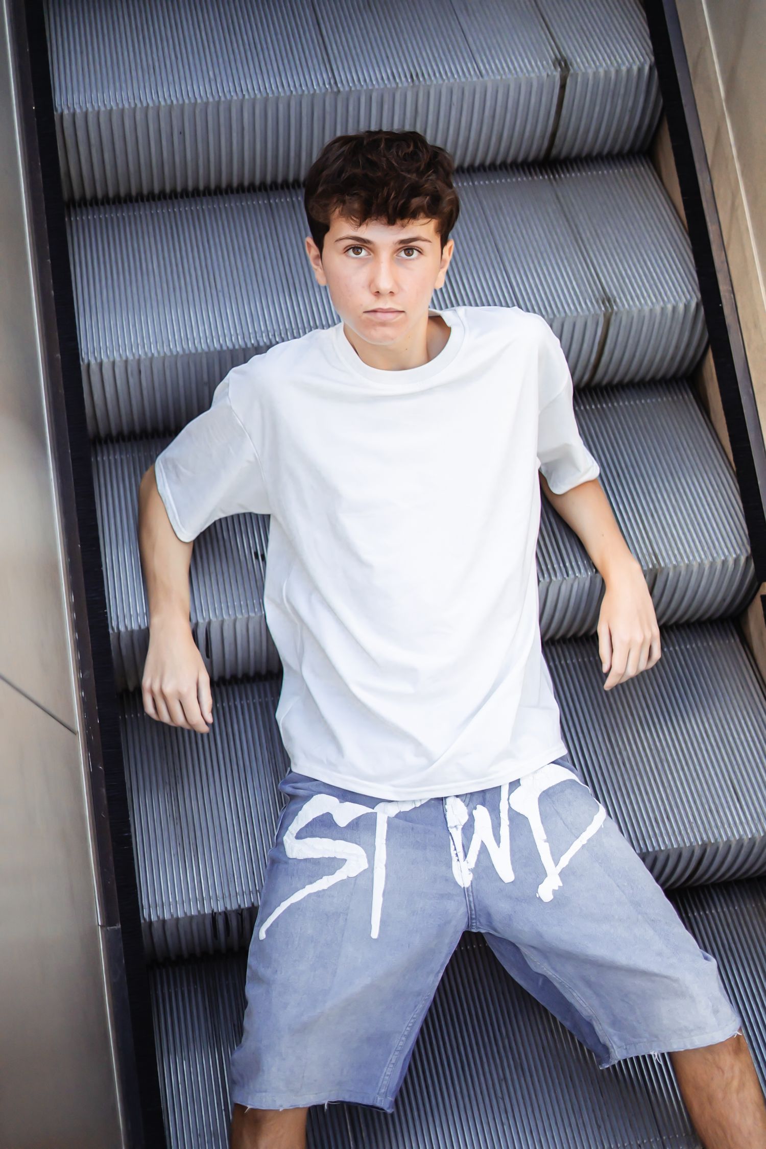 Person in white t-shirt and gray shorts posing on escalator steps in casual urban style.