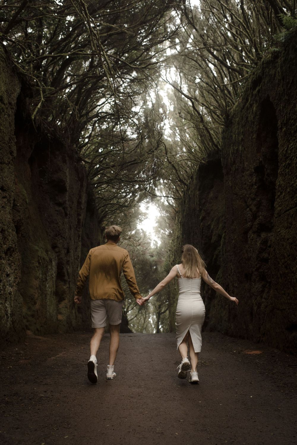 A couple walks hand in hand down a mystical tree-lined path with intertwining branches creating a natural tunnel.
