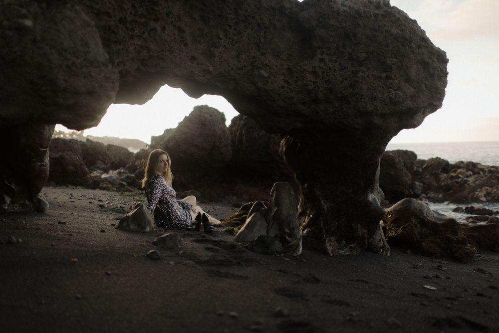 People relax under a dramatic rock arch formation on a moody black sand beach at sunset.