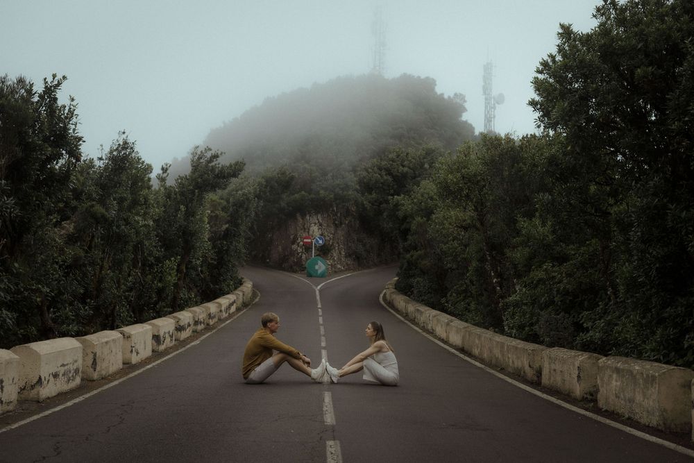 A misty mountain road curves through dense forest with stone barriers along the edges in foggy conditions.